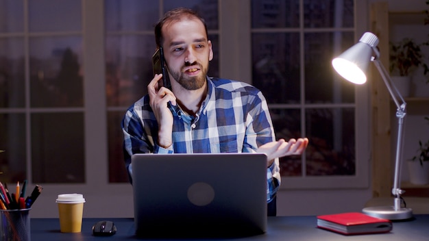 Zakenman die een argumentgesprek voert via de telefoon terwijl hij 's nachts in zijn thuiskantoor werkt.