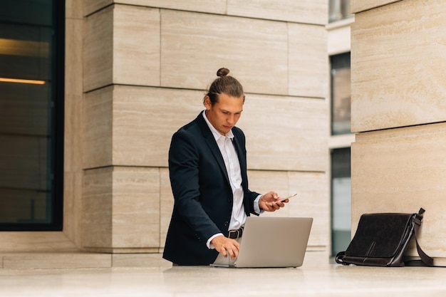 zakenman die een laptop gebruikt terwijl hij een smartphone tegen het gebouw houdt