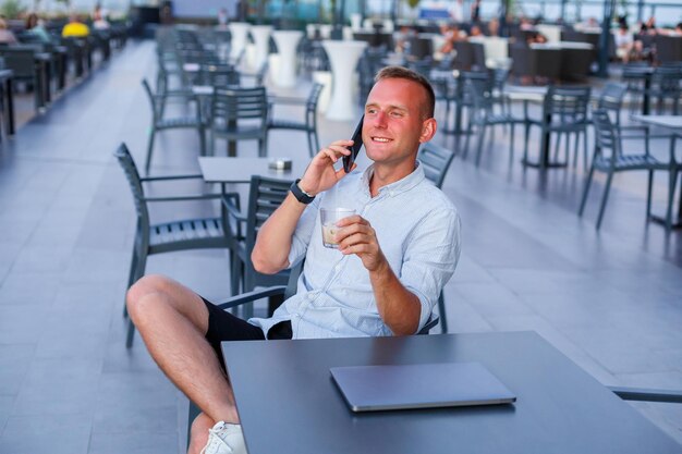 Zakenman die met laptop op het terras van het hotel werkt en aan de telefoon praat om werknemers te beheren tijdens vakantie in het resort Man in korte broek en shirt die een verkoelend drankje drinkt