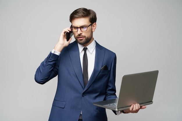 Zakenman die op de telefoon spreekt en laptop houdt die over grijze achtergrond wordt geïsoleerd