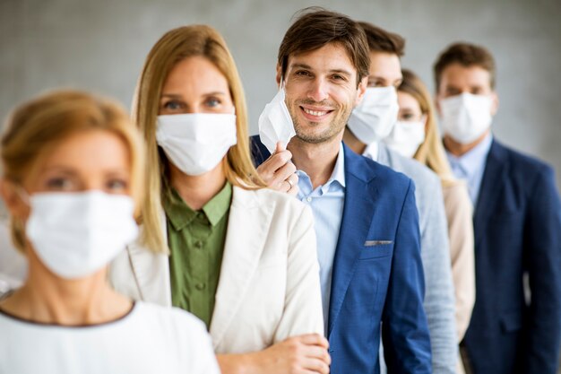 Zakenman die zijn beschermende gezichtsmasker opstijgt en naar de camera kijkt met zijn teamleden die in de rij op kantoor staan