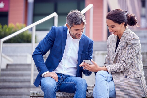 Zakenman en zakenvrouw die stapsgewijs zitten terwijl ze buiten vergaderen en naar de telefoon kijken