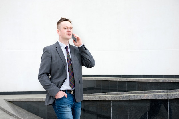 Zakenman, gekleed in een grijze jas, blauwe spijkerbroek, wit overhemd en stropdas komt uit het zakencentrum en praat tijdens het lopen aan de telefoon