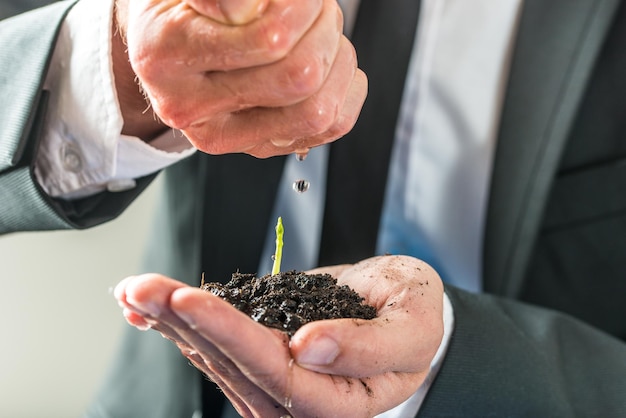 Zakenman gieten water over een kleine groene plant die groeit in vruchtbare grond gehouden in zijn holle linkerhand concept van ecologie milieu en milieuvriendelijke zakelijke close-up