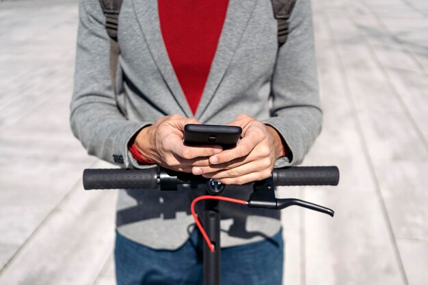 Zakenman in jas met rugzak die op een elektrische scooter staat en een smartphone gebruikt