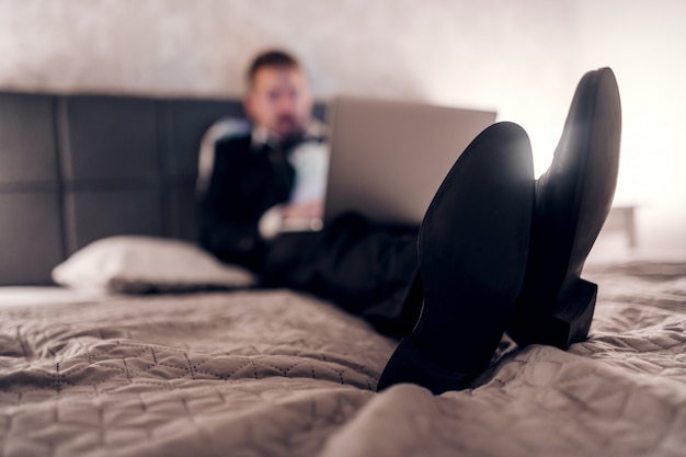 Zakenman in pak zittend op het bed in de hotelkamer en laptop gebruikt voor werk. Overwerk concept. Selectieve aandacht voor schoenen.