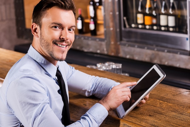 Zakenman in staaf. knappe jonge man in overhemd en stropdas die op digitale tablet werkt en glimlacht terwijl hij aan de bar zit