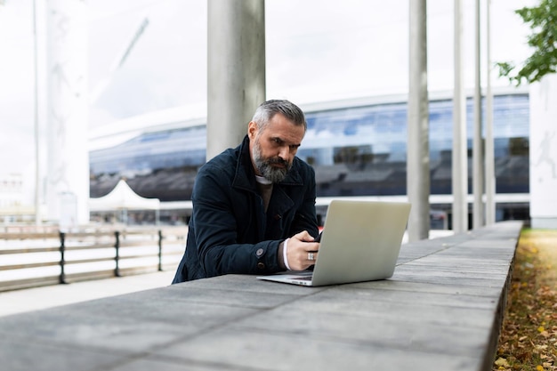 Zakenman man met een grijze baard werkt op een laptop in een stadspark tegen de achtergrond van een kantoor