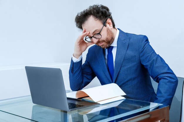 Foto zakenman met doordachte gezicht in blauw pak met behulp van laptop op kantoor interieur achtergrond. concept van het komen met een nieuwe strategie, anticrisisplan