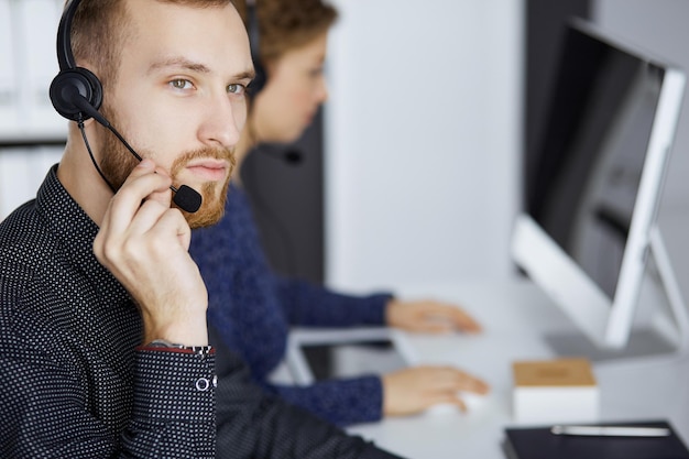 Zakenman met een rode baard praat via de headset in de buurt van zijn vrouwelijke collega terwijl hij in een modern kantoor zit. Diverse mensen groeperen in callcenter. Telemarketing en klantenservice.