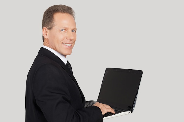 Zakenman met laptop. Achteraanzicht van een senior man in formalwear die iets op laptop typt en over de schouder kijkt terwijl hij tegen een grijze achtergrond staat
