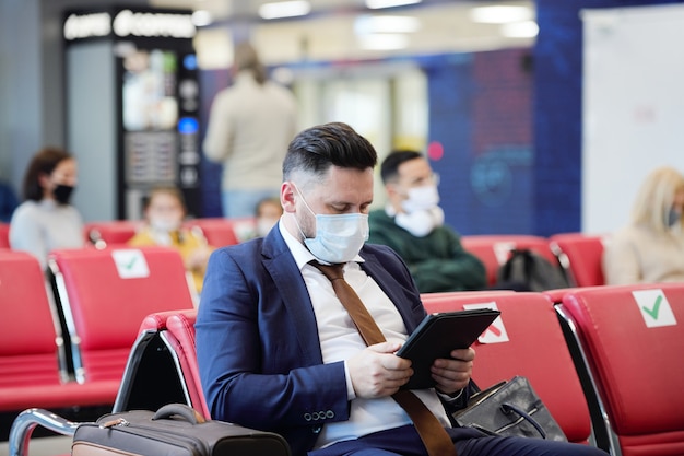 Zakenman met masker die digitale tablet gebruikt terwijl hij in de wachtkamer op de luchthaven zit