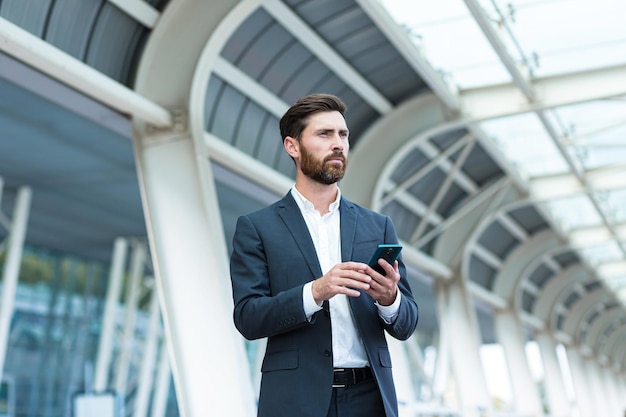 Zakenman staande achtergrond een moderne treinstation luchthaven of bushalte in formeel pak met een koffer met behulp van app mobiele telefoon Reizende zakenman toerist met een smartphone in handen bestel een taxi