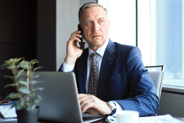 Zakenman van middelbare leeftijd met laptop praten over de telefoon in moderne kantoren.