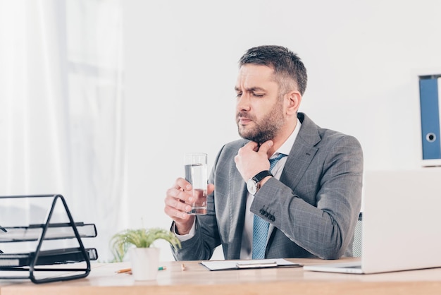 Zakenman zit aan bureau keel aan te raken en glas water vast te houden in kantoor