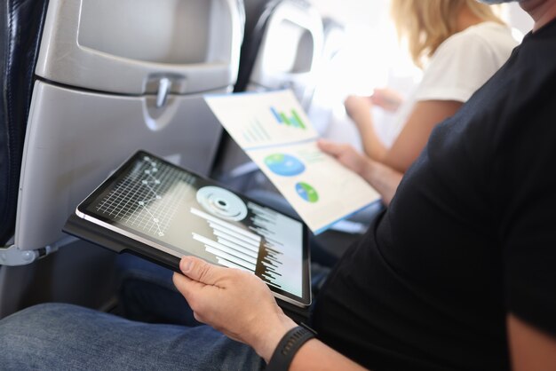 Zakenman zittend in cabine van vliegtuig bestudeert zakelijke grafieken in tablet en documenten