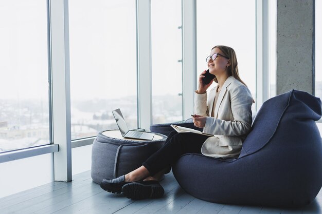 Zakenvrouw denkt tijdens het schrijven in een notitieboekje op het werk Moderne succesvolle vrouw Jong lachend meisje in glazen zit in een stoel aan een tafel in een open kantoor