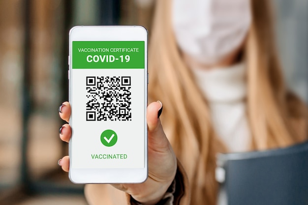 Zakenvrouw die een medisch masker draagt met een scherm van een mobiele telefoon met een applicatie over vaccinatie tegen coronavirus, covid-19 tegen de achtergrond van een gebouw