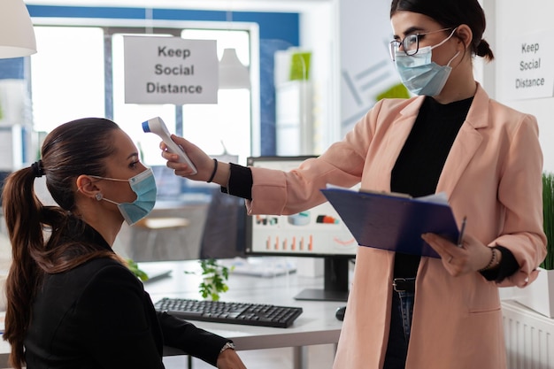 Zakenvrouw die temperatuur meet met behulp van een infraroodthermometer om infectie met coronavirus te voorkomen. Manager met medisch gezichtsmasker tegen covid19 die in bedrijfsbureau werkt tijdens viruspandemie