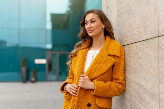 Zakenvrouw gekleed gele jas permanent buitenshuis corporatieve gebouw oppervlak Kaukasische vrouwelijke ondernemer op straat in de buurt van kantoorgebouw met ramen in de stad Stijlvolle zakenvrouw