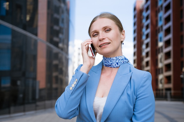 Zakenvrouw in een blauwe jas en jurk praten aan de telefoon met een map met papieren in haar hand
