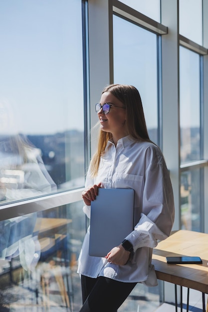 Zakenvrouw in een wit overhemd en bril met een laptop in haar handen op kantoor bij het raam Moderne zakenvrouw in stijlvolle kleding Freelance meisje dat buiten kantoor werkt