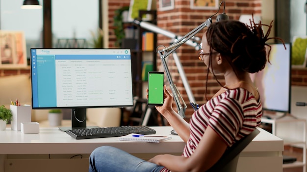 Zakenvrouw kijken naar greenscreen-display op mobiele telefoon, op afstand werk aan het bureau. Werken met geïsoleerde mockup-sjabloon, lege chroma key-weergave en copyspace-achtergrond. Statief geschoten.