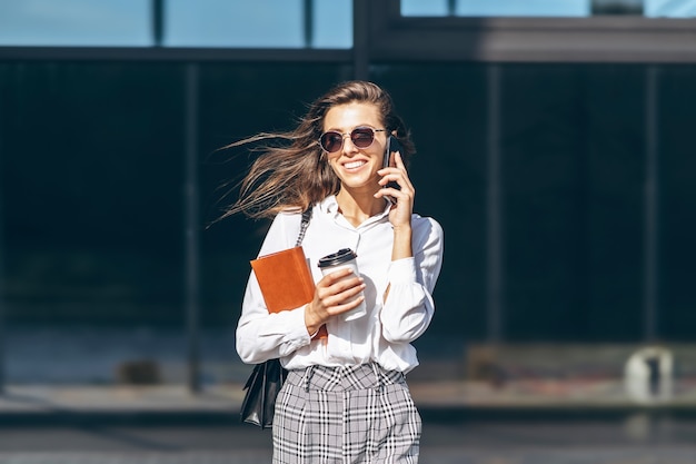 Zakenvrouw lopend en pratend op de mobiele telefoon buiten in de buurt van modern zakencentrum met notebook.