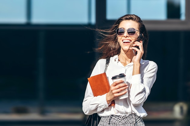 Zakenvrouw lopend en pratend op de mobiele telefoon buiten in de buurt van modern zakencentrum met notebook.