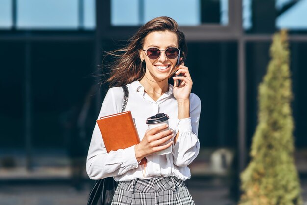 Zakenvrouw lopend en pratend op de mobiele telefoon buiten in de buurt van modern zakencentrum met notebook