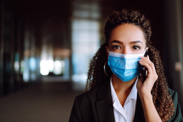 Zakenvrouw met beschermend medisch masker staat in de buurt van kantoor en gebruikt telefoon covid19