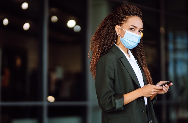 Zakenvrouw met beschermend medisch masker staat in de buurt van kantoor en gebruikt telefoon covid19