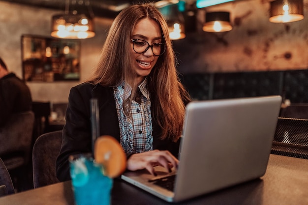 Zakenvrouw met een bril die in een modern café zit en op haar laptop werkt terwijl ze een cocktail drinkt Selectieve focus