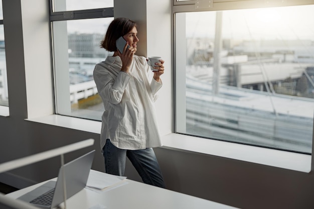 Zakenvrouw met een kopje koffie en pratende telefoon terwijl ze bij het raam op kantoor staat