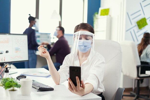 Zakenvrouw met een zakelijke videoconferentie met behulp van smartphone met gezichtsmasker. collega's die werken met respect voor sociale afstand tijdens wereldwijde pandemie met coronavirus.