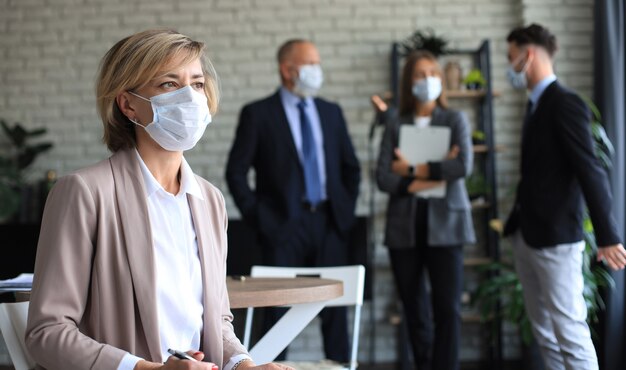 Zakenvrouw met medisch masker met haar personeel, mensengroep op de achtergrond in een modern, licht kantoor binnenshuis.