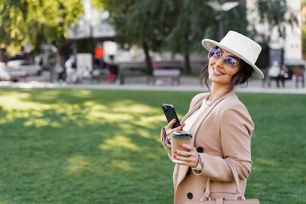 Zakenvrouw met mobiele telefoon en kopje koffie op straat. Aantrekkelijk meisje droeg officiële witte jurk, jas en hoed.