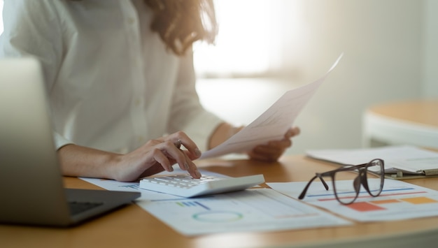 Zakenvrouw of accountant die werkt Financieel Manager Onderzoek Procesboekhouding berekenen met rekenmachine voor het analyseren van marktgrafiekgegevens voorraadinformatieoverzicht op tafel in kantoor.