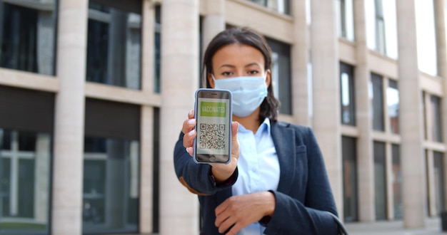 Zakenvrouw van gemengd ras met veiligheidsmasker met smartphonescherm met online vaccinatiepaspoort