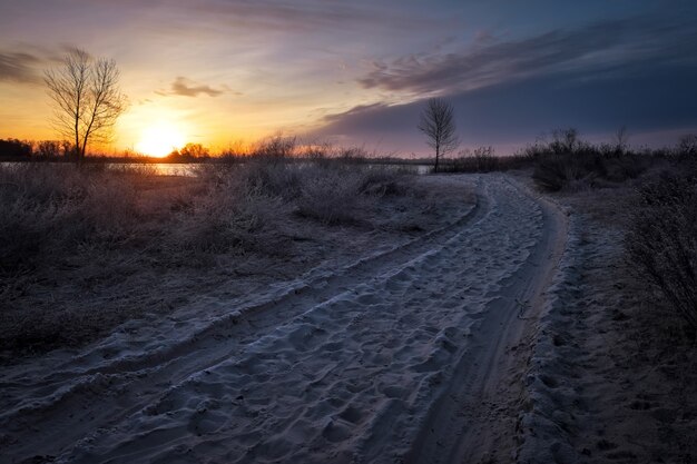 Zandweg en bomen in de vorst, kust van rivier en zon bij zonsondergang.