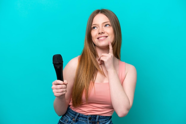 Zanger kaukasische vrouw die een microfoon oppakt die op blauwe achtergrond wordt geïsoleerd en een idee denkt terwijl ze omhoog kijkt