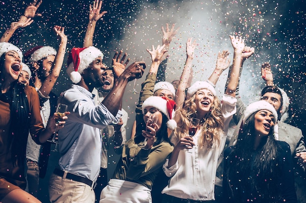 Ze houden van Kerstmis. Groep mooie jonge mensen in kerstmutsen die kleurrijke confetti gooien en er gelukkig uitzien