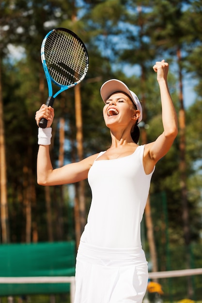 Ze is de beste! Gelukkige jonge vrouw in sportkleding met tennisracket en gebaren terwijl ze op de tennisbaan staat