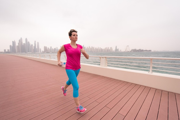 zeer actieve jonge mooie vrouw bezig met rennen op de promenade langs de oceaanzijde met een grote moderne stad op de achtergrond om haar fitnessniveaus zoveel mogelijk op peil te houden