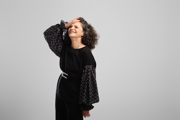 Zeer gelukkige senior vrouw met weelderig krullend haar lachen in studio over grijze achtergrond