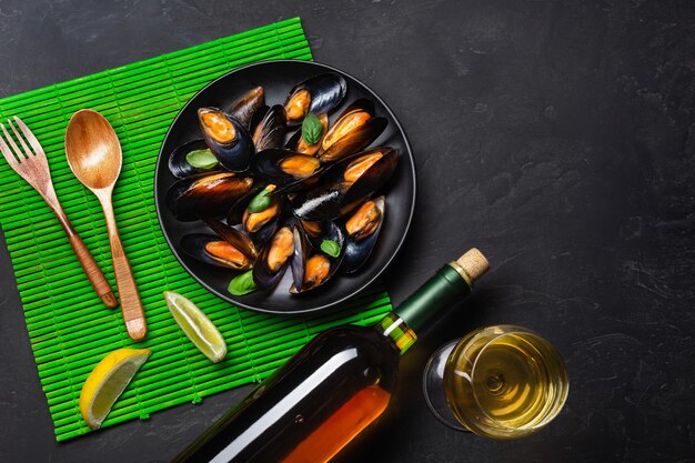 Zeevruchtenmosselen, basilicumbladeren in een zwarte plaat met wijnbootle, wijnglas, citroen, houten lepel en vork op groene bamboemat en stenen tafel. Bovenaanzicht.