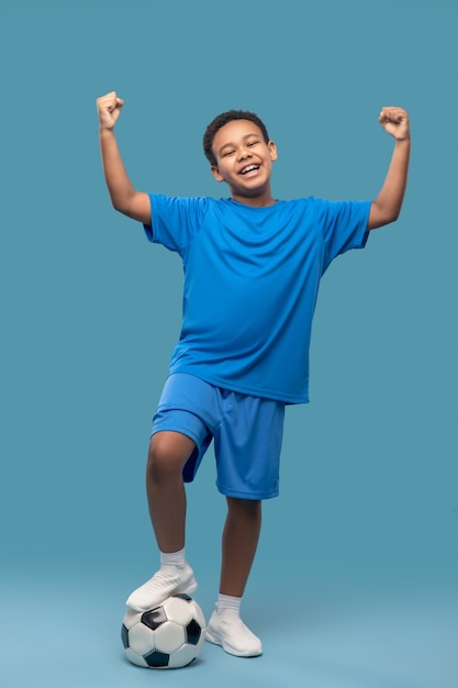 Zege. Gelukkige jongen met donkere huid in blauwe sportkleding met voetbal die zijn handen zegevierend opheft