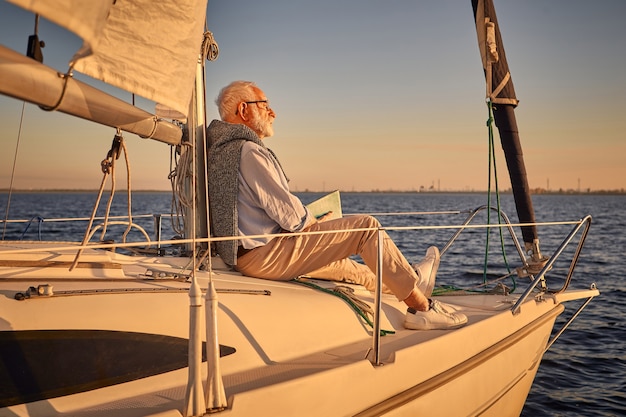 Zeilende man senior man zit aan de zijkant van zijn zeilboot of jacht drijvend in de zee lezen