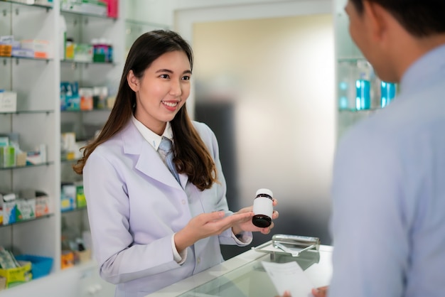 Zekere Aziatische jonge vrouwelijke apotheker met een mooie vriendelijke glimlach en uitleggen van medicijnen aan haar klant in de apotheek. Geneeskunde, farmacie, gezondheidszorg en mensenconcept.