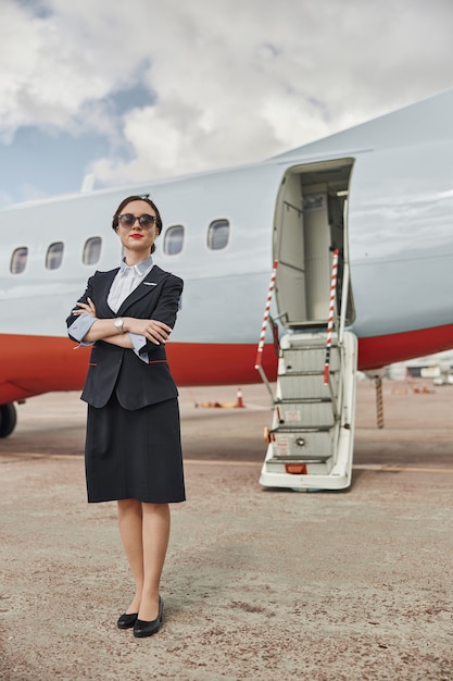 Zekere stewardess op baan dichtbij vliegtuigstraal. modern passagiersvliegtuig met ladder. jonge vrouw met gekruiste armen dragen uniform en bril. civiele commerciële luchtvaart. vliegreisconcept